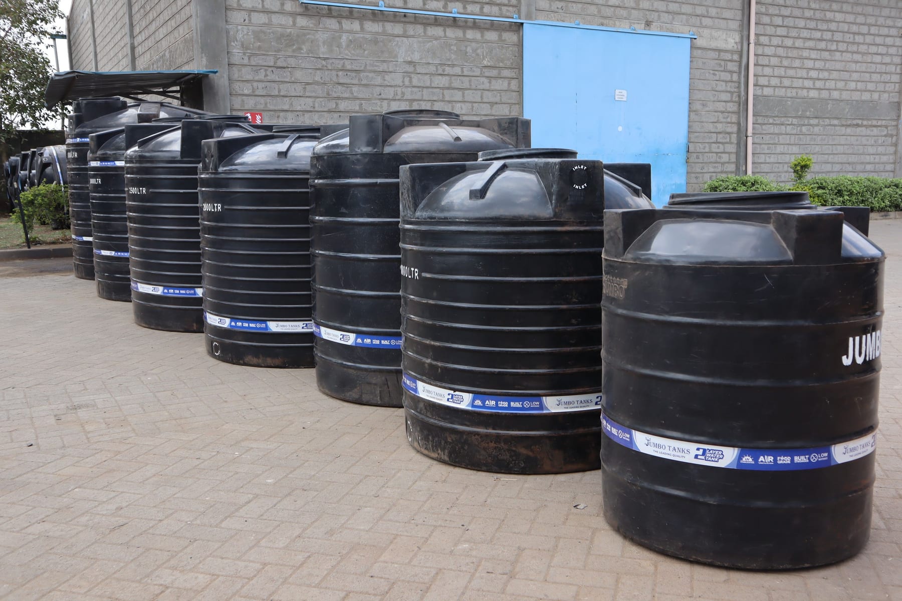 A curved row of black Jumbo cylindrical tanks in the factory yard, showing 2,500, 2,000 and 3,000 litre capacities.